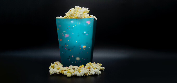 Ein bedruckter Popcornbecher ist vor einem schwarzen Hintergrund in Szene gesetzt. Aus dem personalisierten Popcornbecher ragt etwas Popcorn heraus. Und auch vor dem Individuellen Popcornbecher liegt Popcorn.